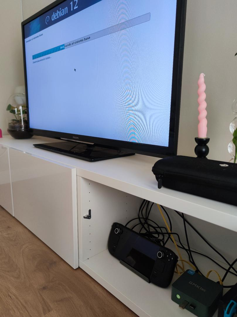 Imatge on es veu un moble de televisió blanc amb tres calaixos, un d’ells obert on es mostra una Steam Deck en un dock, el mini pc GMKtec g3 i la Nintendo Switch. A sobre del moble hi ha una televisió on es mostra en pantalla el procés d’instal·lació de debian 12